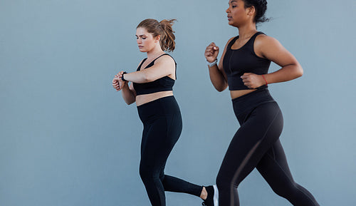 Two female friends running against a grey wall. Plus size female checking her smartwatch while jogging outdoors with her friend.