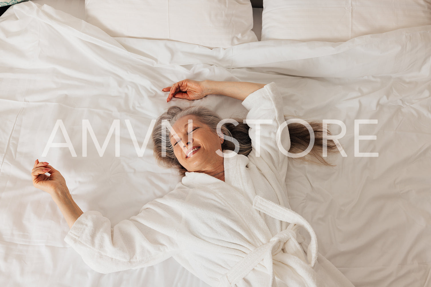 Happy senior woman in bathrobe lying on a bed in hotel. Cheerful aged female enjoying morning.