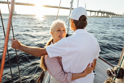 Smiling female looks over the shoulder of her husband and hugging him. Affectionate couple standing together on sailboat.