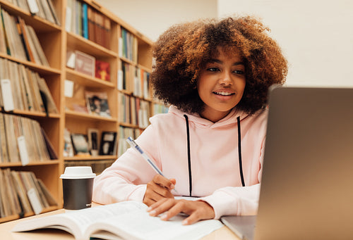 Smiling girl in hooded sweatshirt looking at laptop while sitting in library