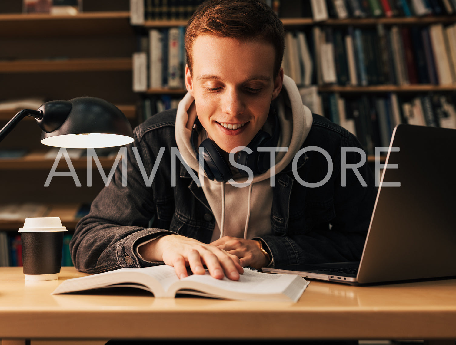 Smiling teenager reading from book. Student with laptop and a book sitting at desk in library.