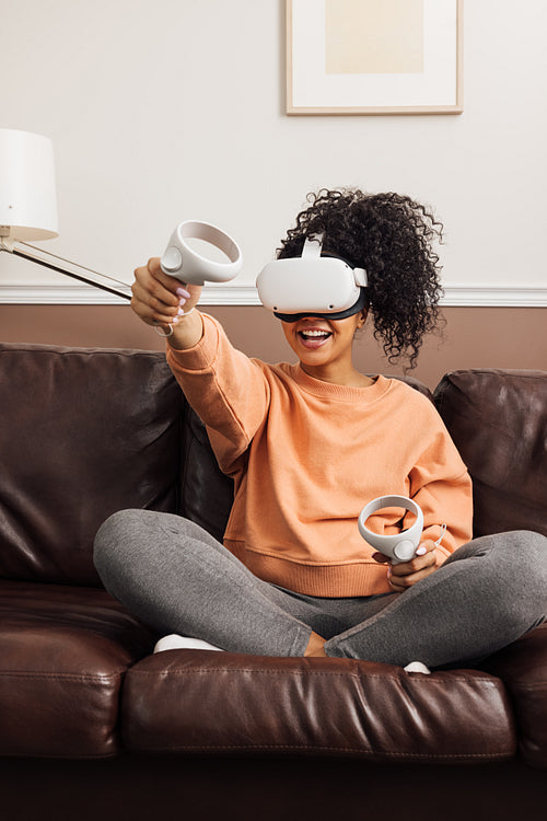 Woman with joysticks and VR goggles playing video games on a sofa