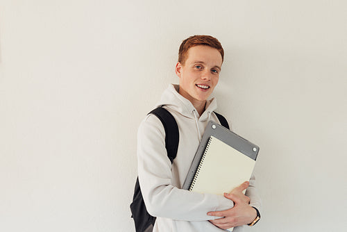 Student with laptop standing at wall and looking at camera. Smiling male in casuals leaning a wall.