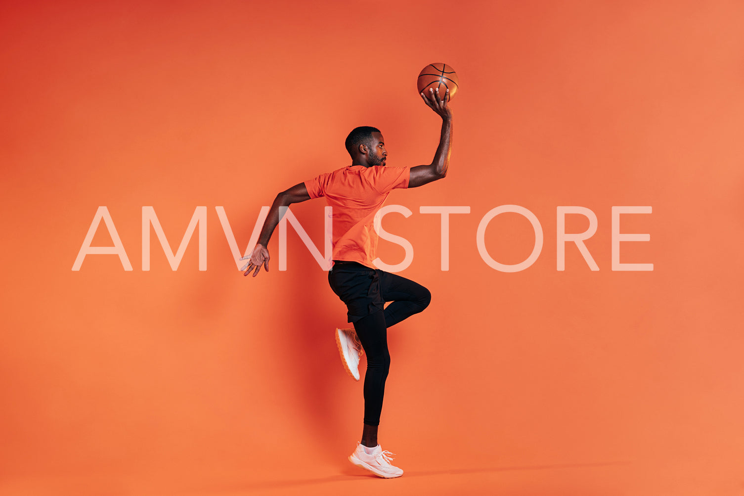 Side view of sportsman jumping in the air with basket ball in hand. Male in sportswear exercising indoors at an orange wall.