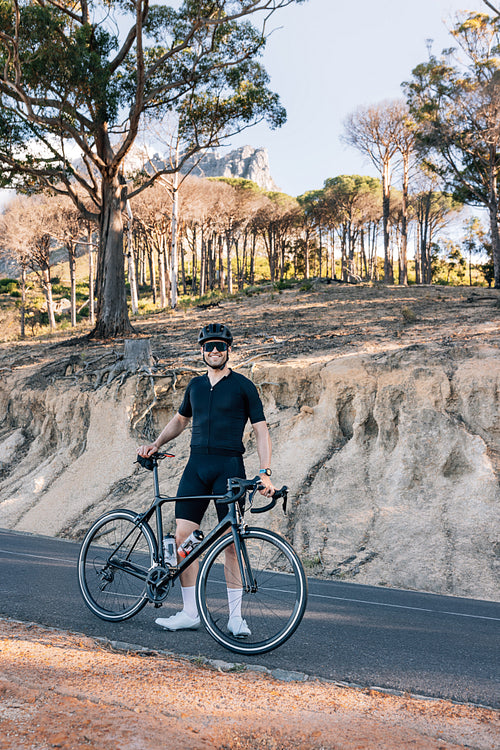 Full length of a smiling triathlete with bicycle standing on an empty road and looking at camera