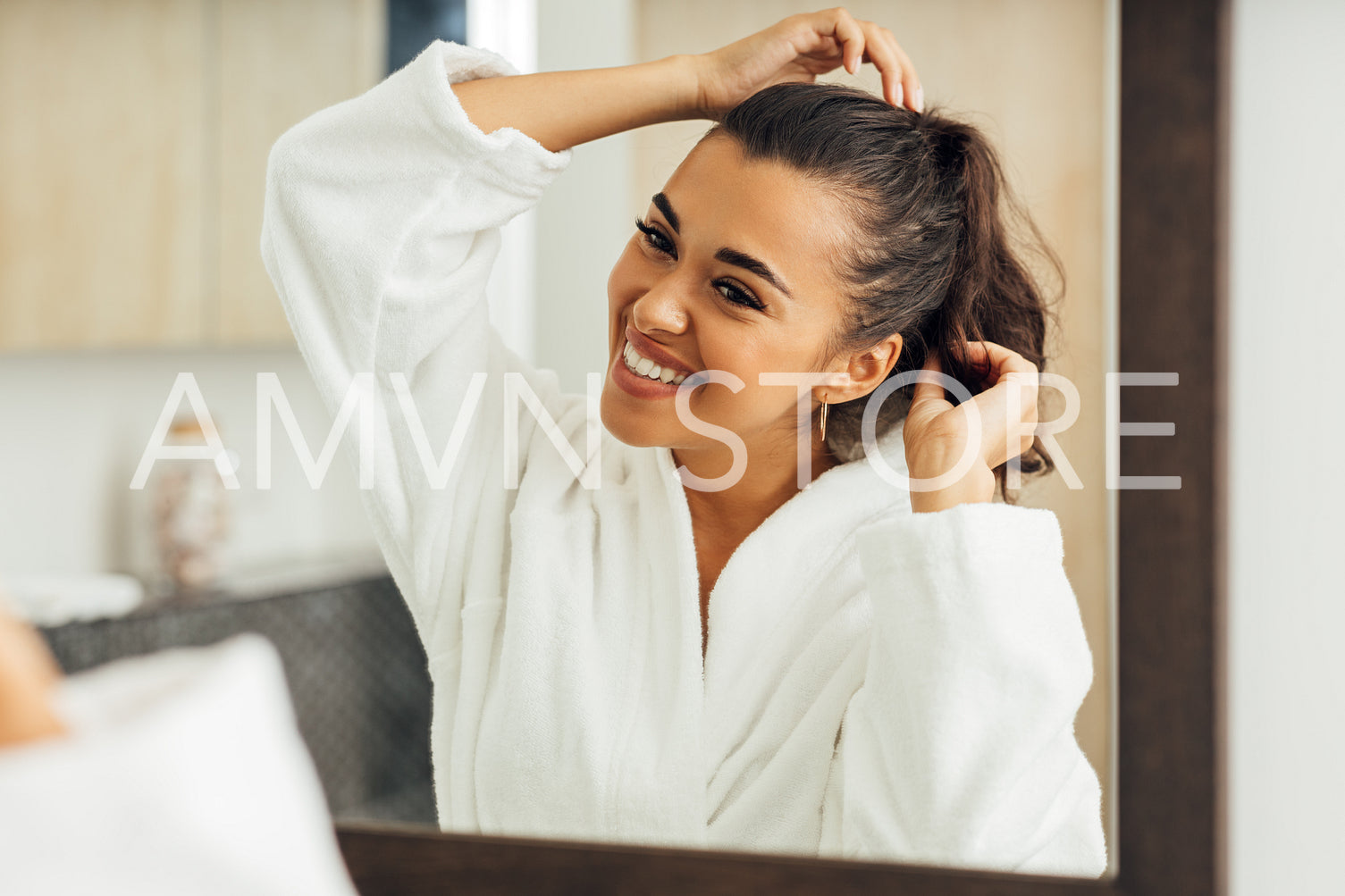 Beautiful smiling woman wearing bathrobe looking at mirror and adjusting her hair	