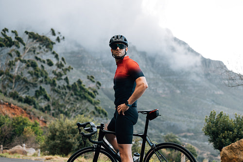 Male cyclist relaxing during his outdoor ride. Cyclist wearing helmet and glasses with bike against mountain looking away.