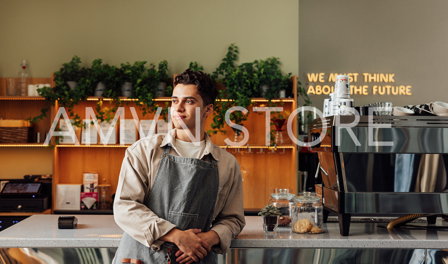 Confident local coffee shop owner leaning counter wearing an apron. Male barista looking away in a cafe.