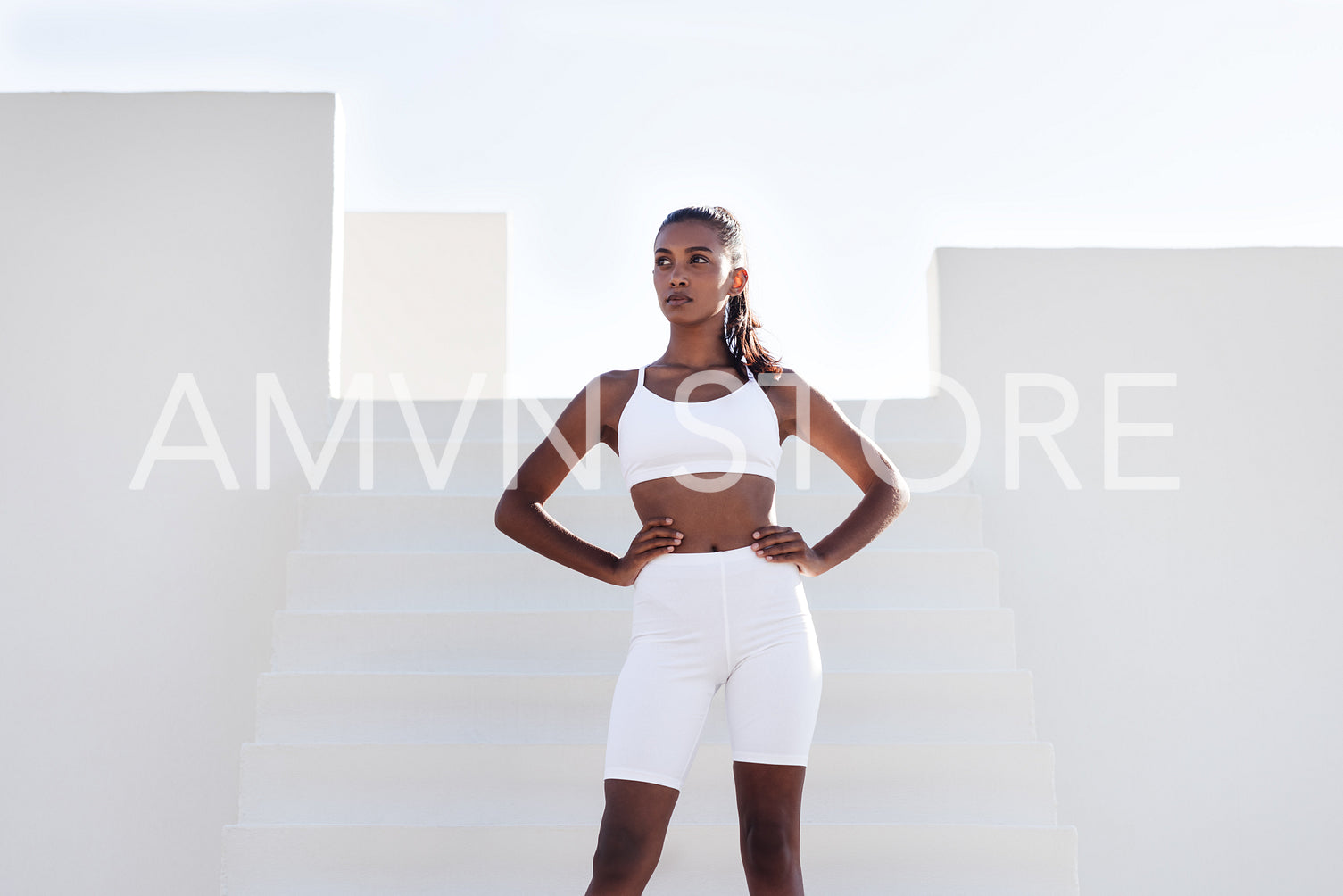 Confident and fit woman standing on the stairs and looking away. Slim female in white sports wear taking a break with hands on her hips.