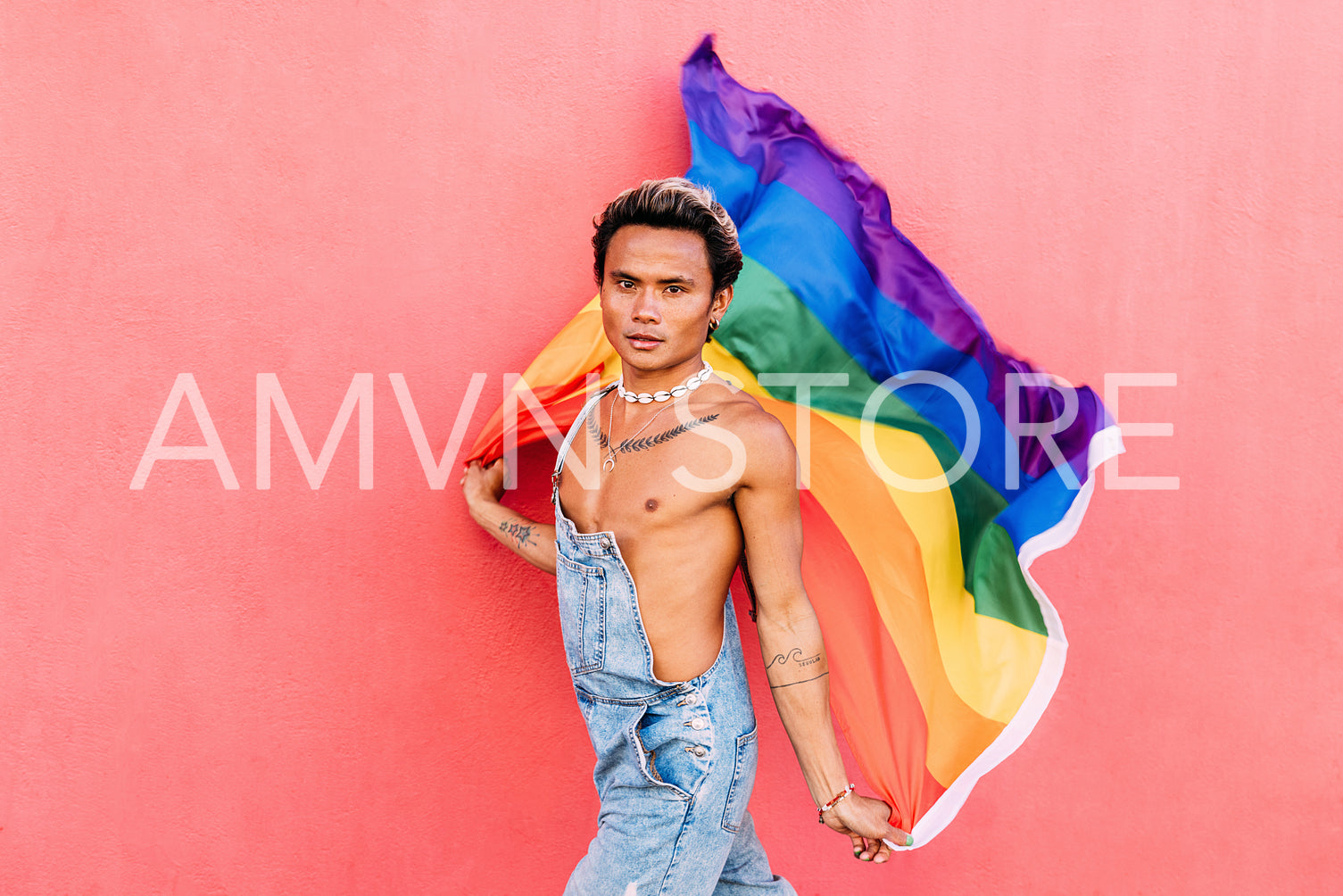 Portrait of a handsome young man holding pride movement LGBT rainbow flag against a pink wall. Man with a pride flag looking at camera outdoors.