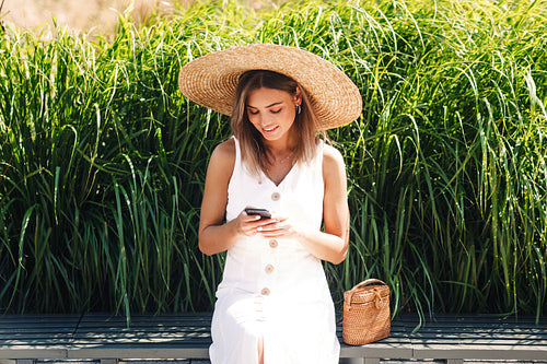 Stylish woman wearing straw hat and using mobile phone while sitting in park