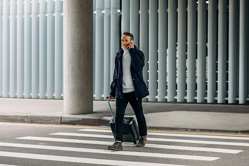 Senior man talking on cell phone while walking on pedestrian crosswalk and pulling suitcase