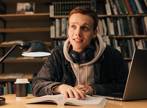 Smiling student sitting in front of bookshelf and reading. Guy in casuals looking away in library at evening.