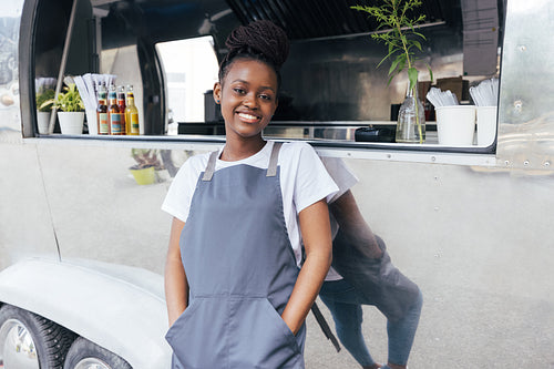 Portrait of smiling female owner in apron leaning on a food truck and looking at camera