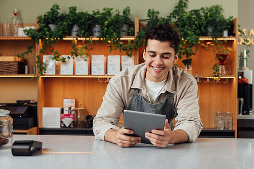 Smiling coffee shop owner at the counter with digital tablet. Male bartender in an apron leaning counter looking at a portable computer.