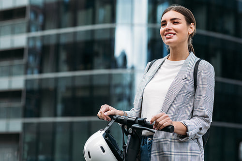 Smiling woman holding a handlebar of electric push scooter while standing at glass building