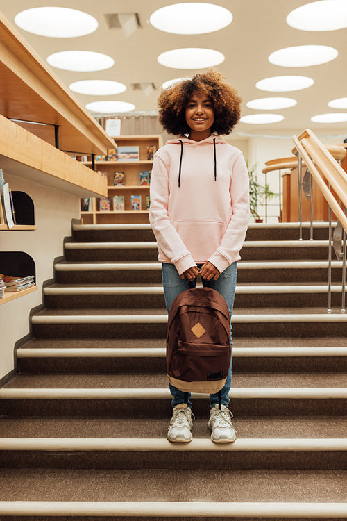 Smiling girl with curly hair holding backpack while standing on stairs in library