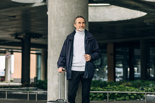 Stylish senior tourist standing outdoors with suitcase and looking away