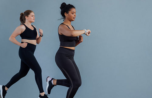 Two females with different body types running outdoors a grey wall. Young woman checking her smartwatches while running outdoors with a fitness buddy.