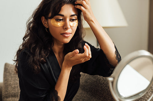 Woman in satin robe looking at mirror and applying face patch in room