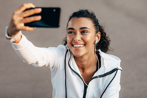 Happy woman sitting on city street wearing headphones and taking selfie after workout