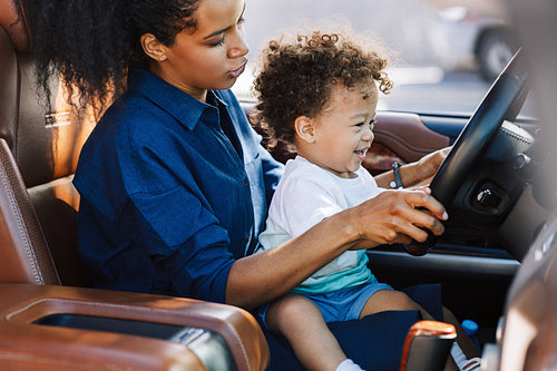 Shot of an adorable little boy sitting on his mother's lap and holding steering wheel. Mother playing with her son in car.