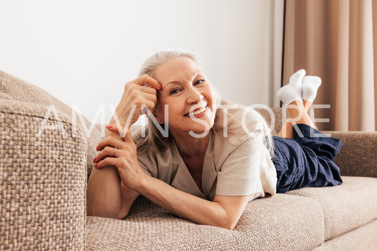 Portrait of a smiling aged woman with grey hair lying on a couch in living room looking at camera