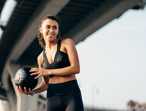 Smiling fit woman holding a medicine ball under a highway and looking away