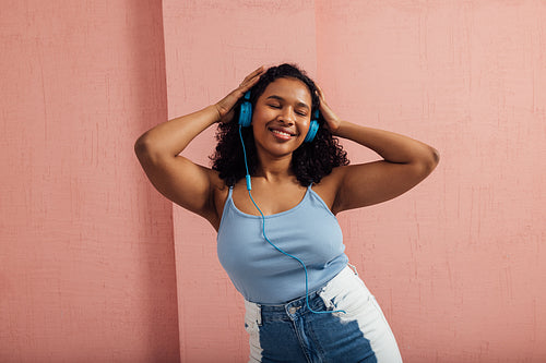 Plus size woman wearing blue headphones and listening to music with closed eyes against a pink wall