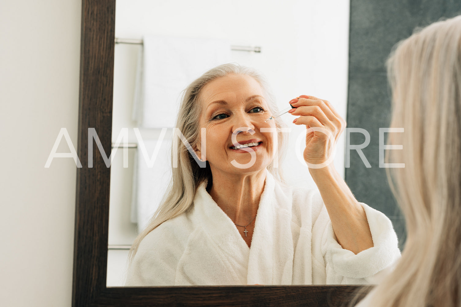 Smiling aged female applying the liquid facial treatment on face. Aged female with a hyaluronic dropper in bathroom.