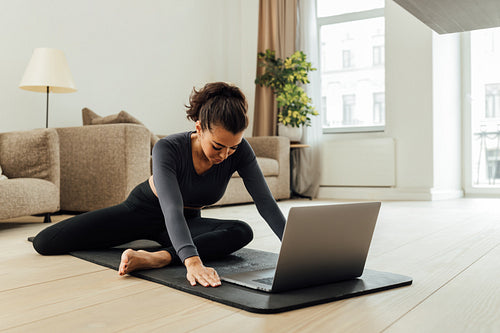 Young middle east woman doing yoga exercise at home. Female in sportswear sitting on mat and stretching her legs in front of a laptop.