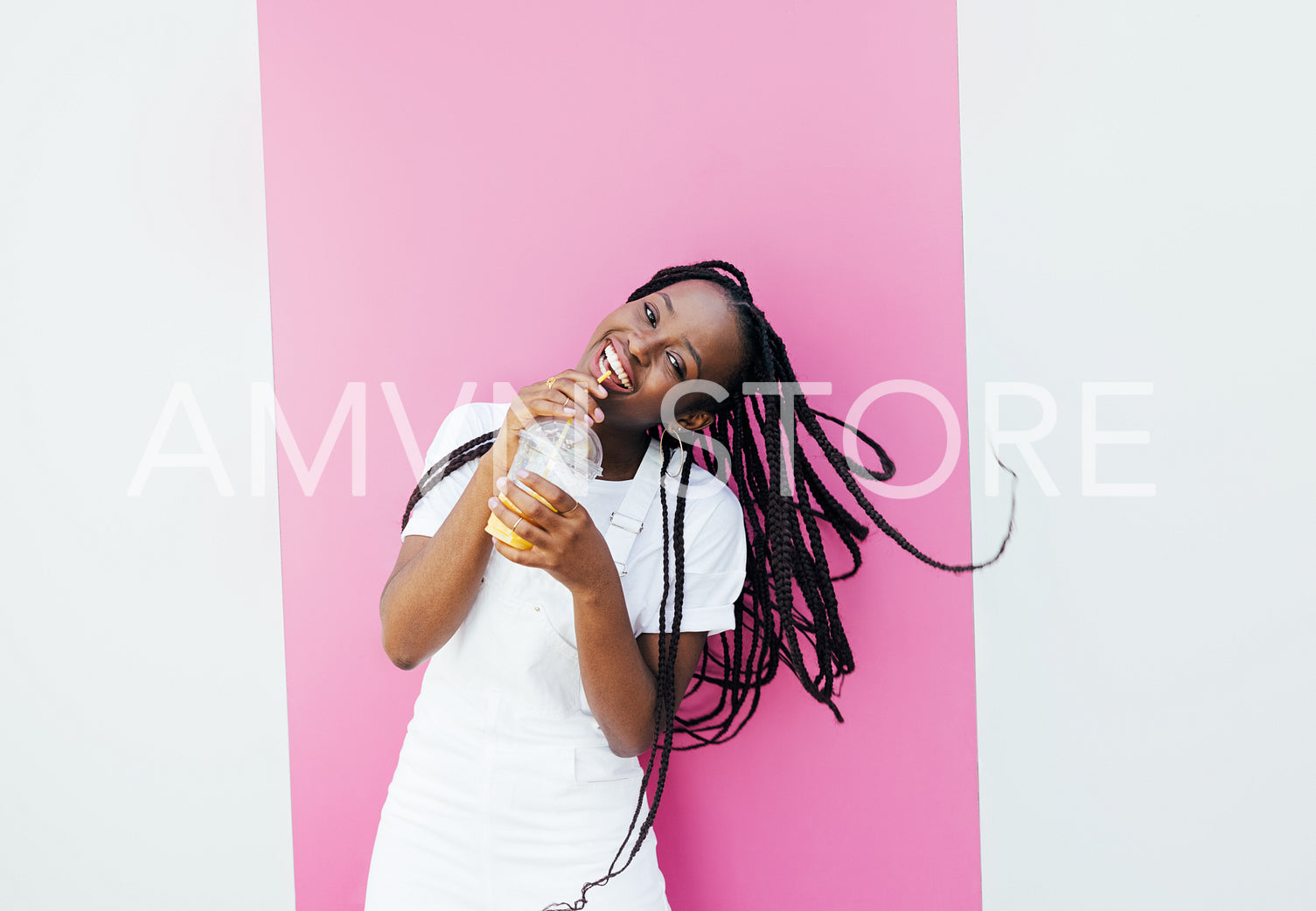 Cheerful woman with braids drinking a juice and looking at camera while standing at pink wall