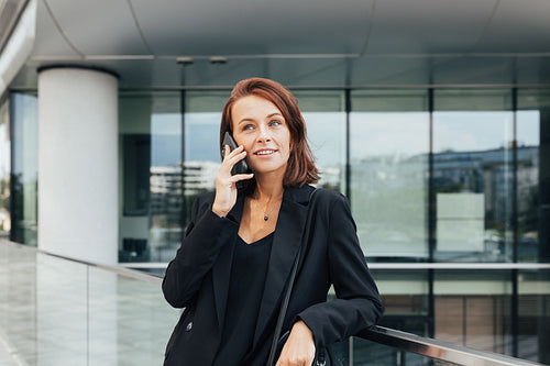 Smiling middle-aged business woman with ginger hair leaning on a railing talking on a mobile phone