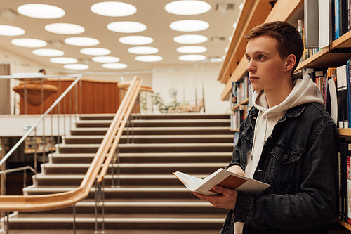 Side view of male student in library holding a book. University student standing at bookshelf.