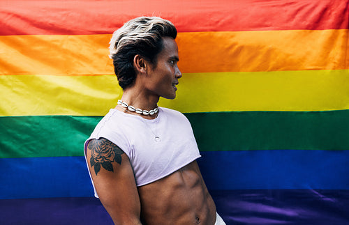 Side view of young stylish guy standing against LGBT flag and looking away