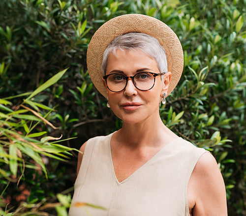 Senior woman with short grey hair, glasses, and straw hat looking at camera in a park