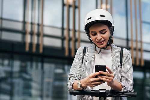 Woman in formal wear typing on smartphone while leaning on the handlebar of electric scooter while standing at an office building