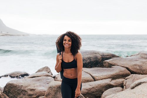 Portrait of a happy woman with yoga mat walking at coastline at sunset