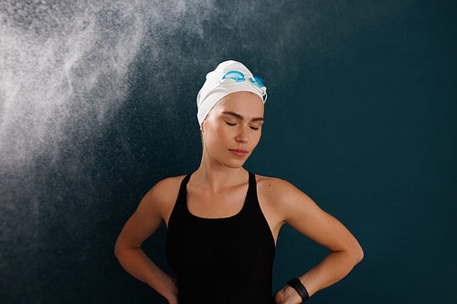 Professional female swimmer wearing a white swimming cap and goggles standing with closed eyes against blue backdrop