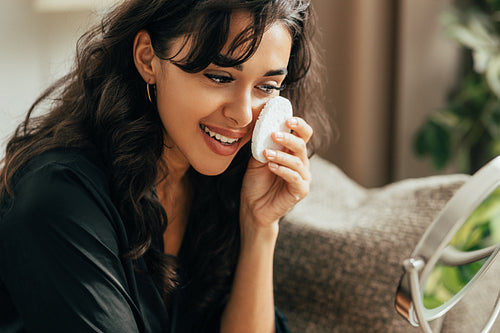 Young brunette woman touching her face with sponge looking at a mirror