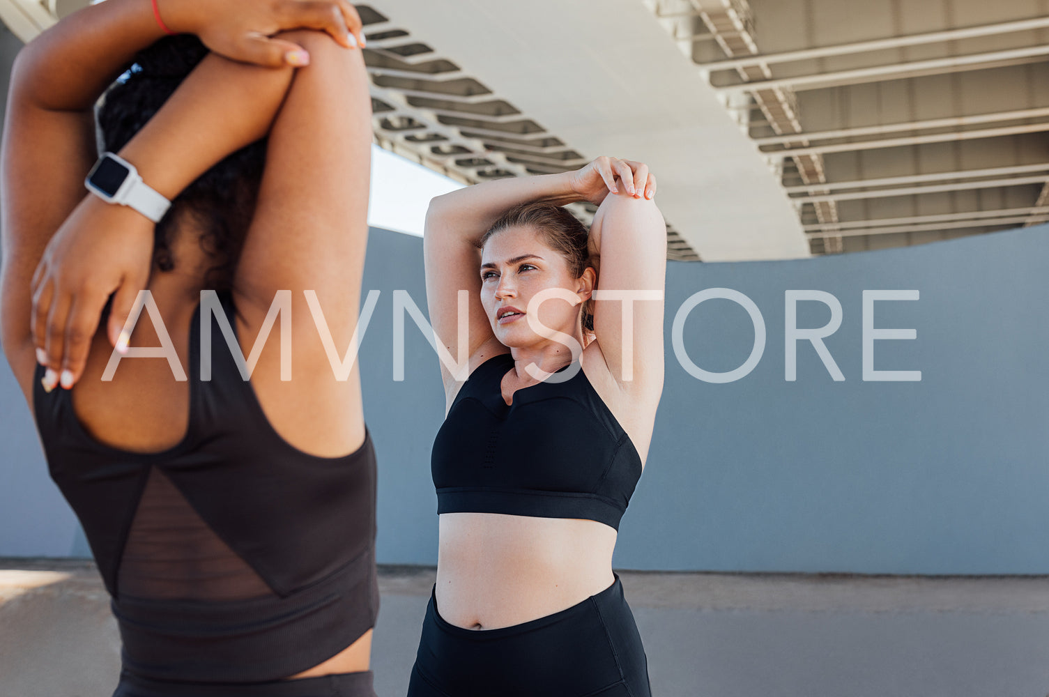 Two plus-size women stretch their hands while standing under a bridge. Young woman warming up hands while standing outdoors with her friend.