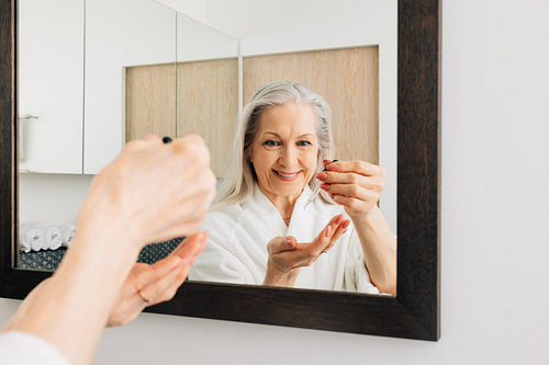 Senior woman in the bathroom doing morning routine. Female holding dropper with hyaluronic acid in front of a mirror.