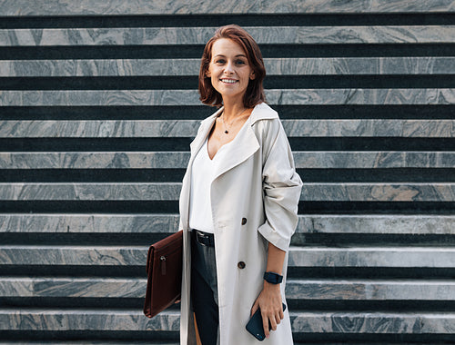 Cheerful middle-aged woman in stylish clothes looking at the camera while standing outdoors. Female in coat with leather folder. Woman with ginger hair wearing stylish clothes.