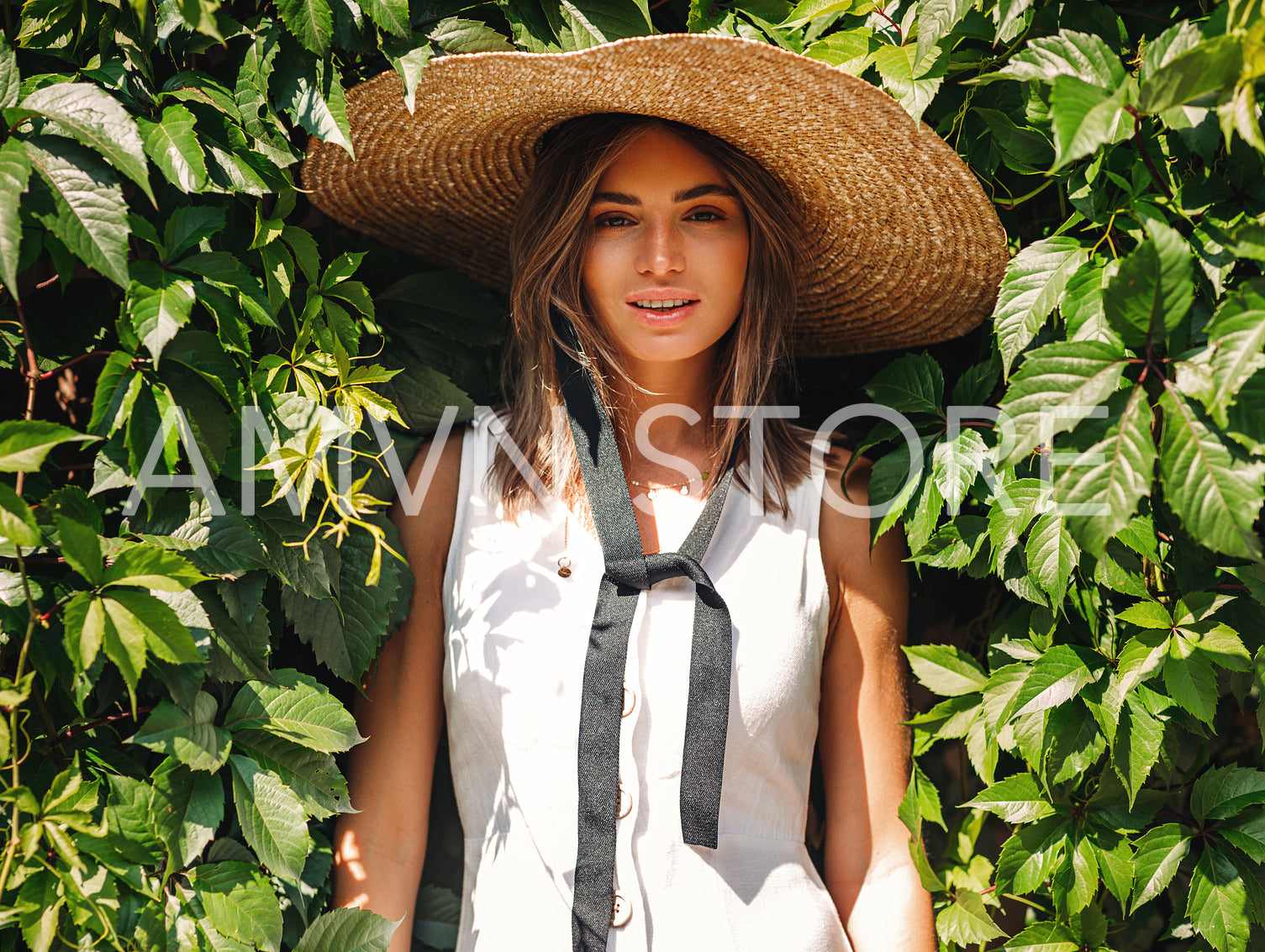 Young blond woman in big staw hat standing at wall with green leafs and looking at camera	