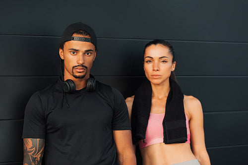 Couple in fitness wear relaxing at a black wall. Man and woman in sportswear looking at camera after a workout.