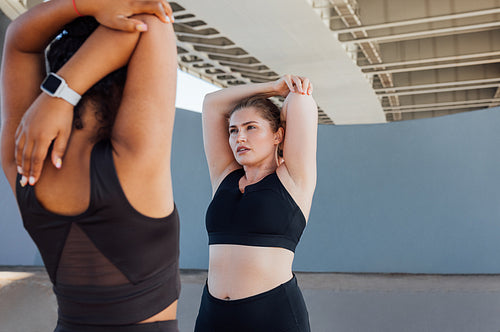 Two plus-size women stretch their hands while standing under a bridge. Young woman warming up hands while standing outdoors with her friend.