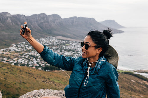 Young woman in sunglasses holding a smartphone and taking a selfie while taking a break during a mountain hike