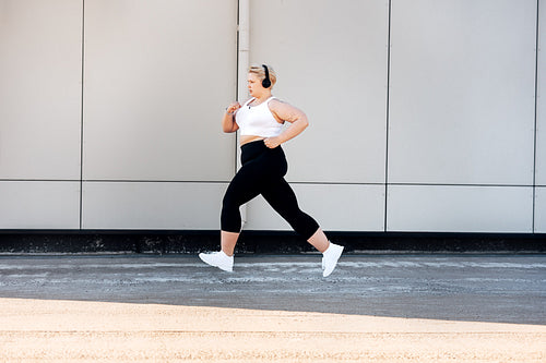 Side view of plus size woman wearing headphones running near a wall. Curvy female in sports clothes jogging outdoors.