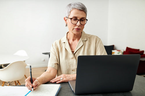 Mature woman looking at laptop screen making notes at home