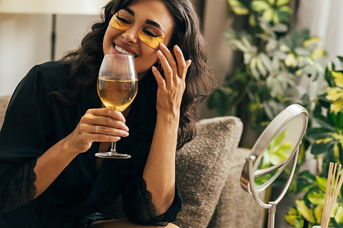 Smiling woman in satin robe relaxing at home. Beautiful female sitting on a couch holding a wineglass adjusting eye patch.